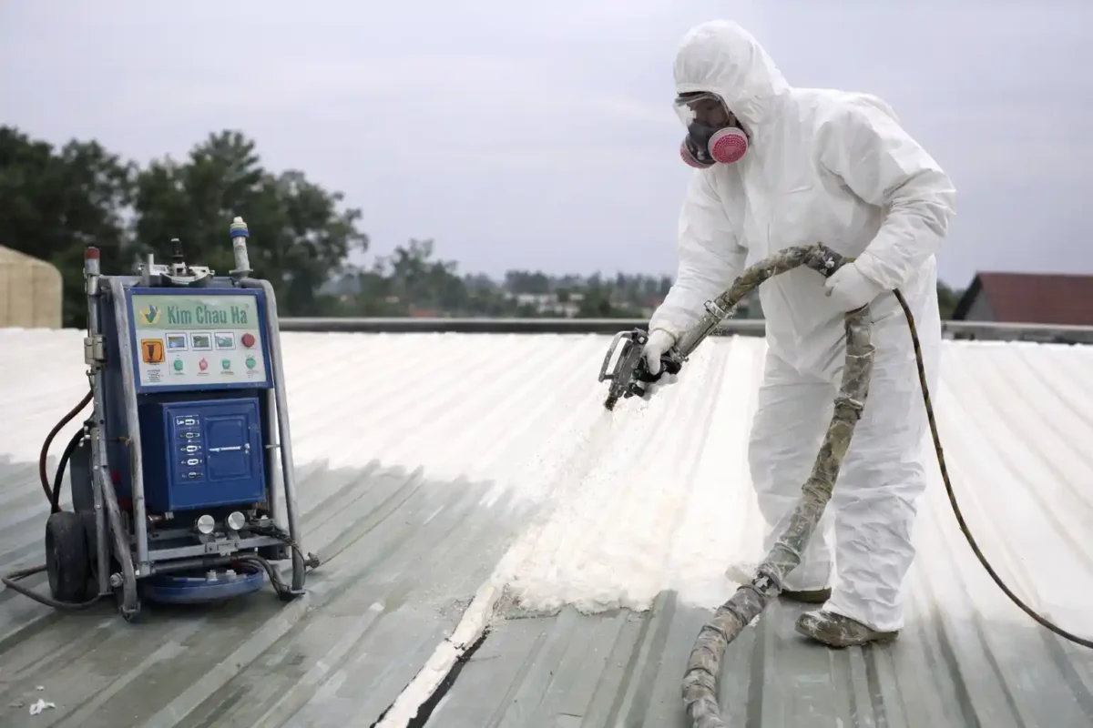 Công nhân phun foam cách nhiệt trên mái tôn tại Quy Nhơn bằng máy phun PU Foam, mặc đồ bảo hộ chống độc đầy đủ.